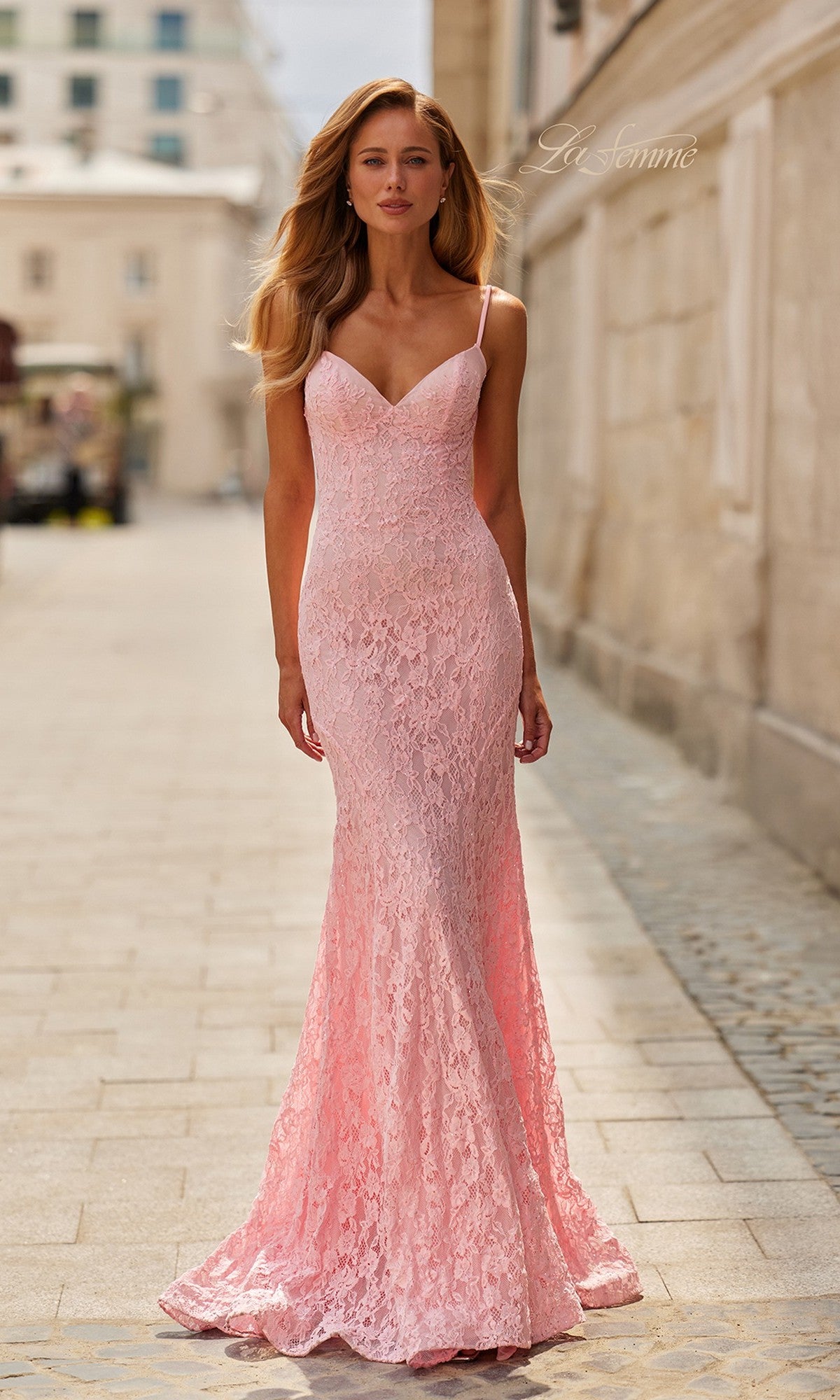 A woman in the La Femme 33452 long prom dress in baby pink, featuring a fitted lace design with thin straps, stands on a stone-paved street beside a light-colored building.